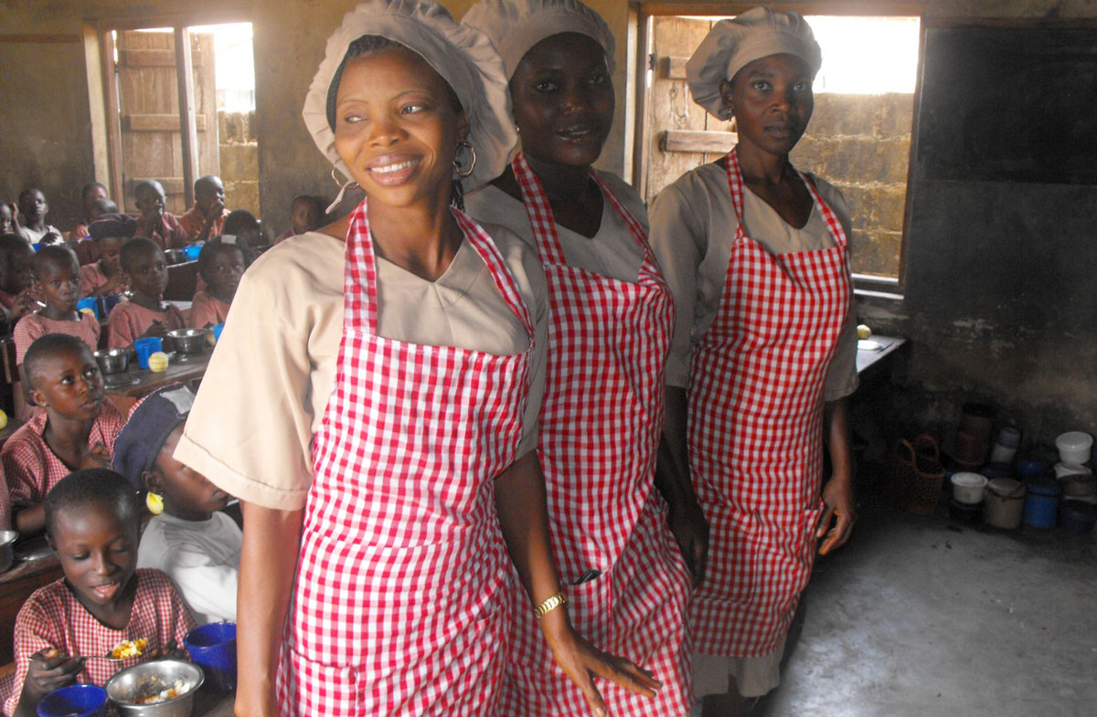 Three women in checkered aprons stand in front of a classroom of children eating from metal bowls.